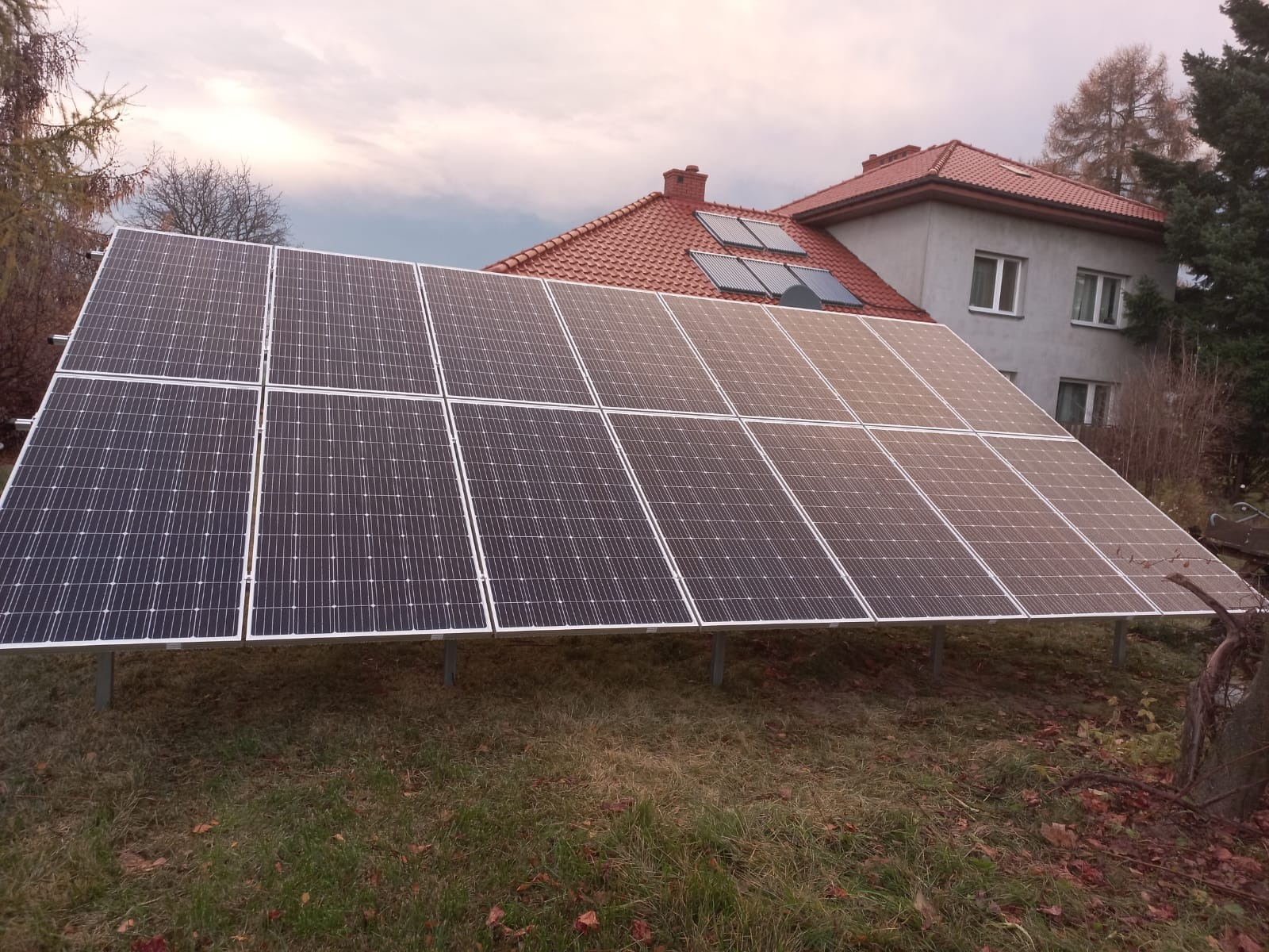 Panele słoneczne zamontowane na ziemi przed domem z czerwonym dachem i kolektorami słonecznymi.