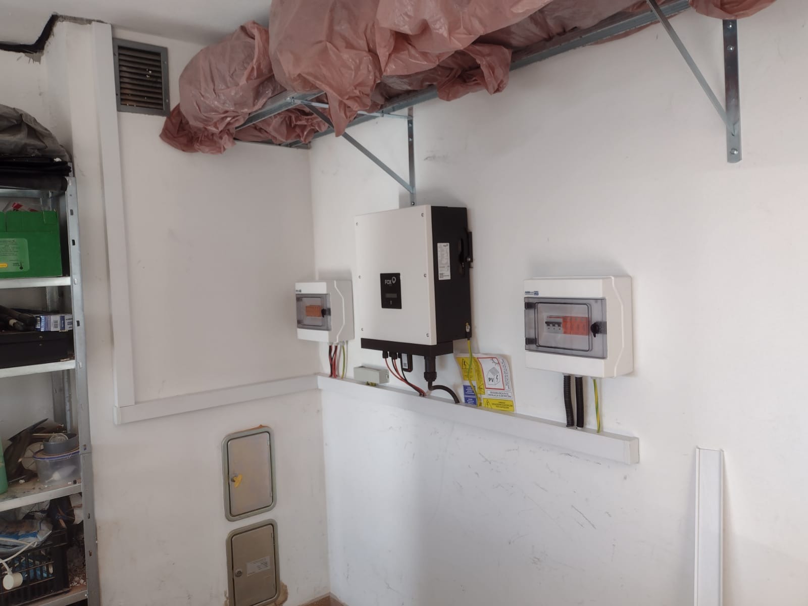 Wnętrze pomieszczenia z zamontowanym inwerterem fotowoltaicznym, dwoma skrzynkami zabezpieczeń oraz instalacją elektryczną. Sufit zabezpieczony folią budowlaną.