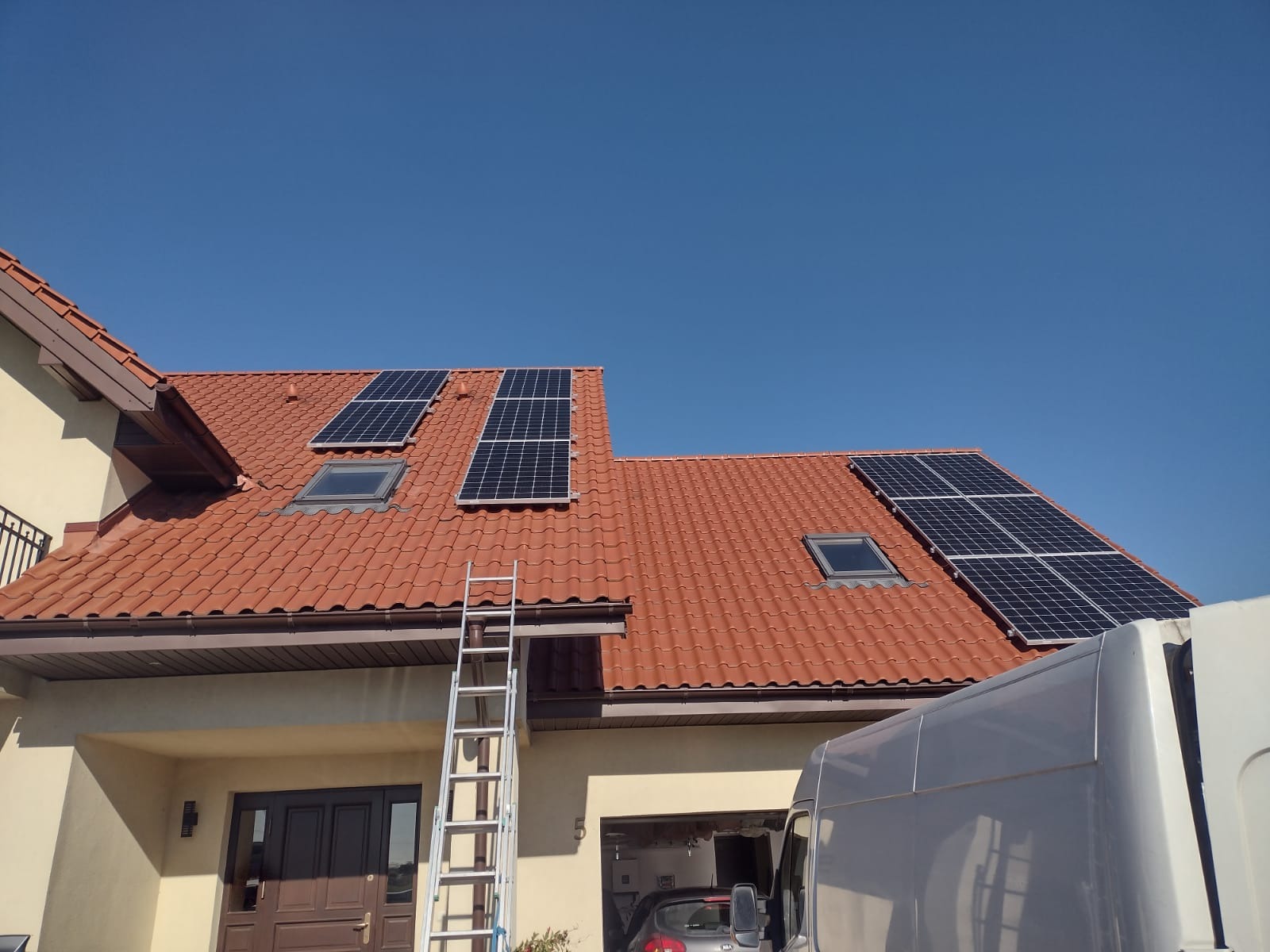 Instalacja paneli słonecznych na dachu domu z czerwoną dachówką, widoczna drabina i częściowo samochód dostawczy.
