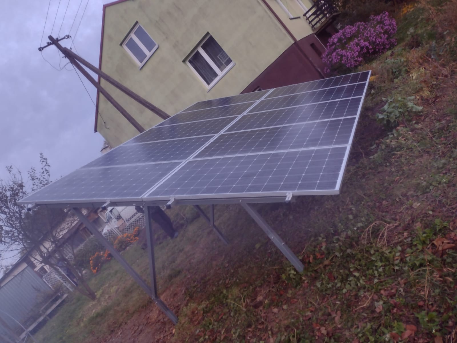 Panel fotowoltaiczny zamontowany na metalowej konstrukcji na tle domu z kwiatami i słupem energetycznym w pochmurny dzień.