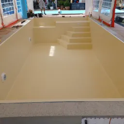 Wnętrze nowo wybudowanego basenu z beżową, gładką powierzchnią, schodami i wbudowanymi lampami, otoczonego betonową konstrukcją i częściowo zadaszonego namiotem.