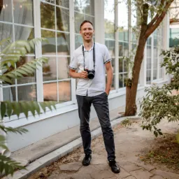 Uśmiechnięty mężczyzna w białej koszuli i ciemnych spodniach, stojący na zewnątrz z aparatem fotograficznym w dłoni, na szyi pasek od aparatu, w tle białe okna i zieleń.