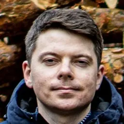 Portret mężczyzny w niebieskiej kurtce na tle stosu drewna, lekko uśmiechnięty, z widocznymi zmarszczkami mimicznymi wokół oczu.