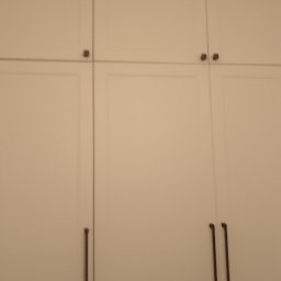 ŚwierG - Biała szafa wnękowa z minimalistycznymi, długimi uchwytami. Górne szafki otwierane na dotyk. Prosty design, jasne wnętrze, pionowy kadr.