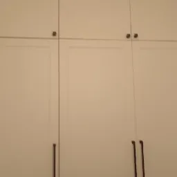 Biała szafa wnękowa z minimalistycznymi, długimi uchwytami. Górne szafki otwierane na dotyk. Prosty design, jasne wnętrze, pionowy kadr.