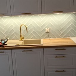 ŚwierG - Biała kuchnia z złotymi akcentami: bateria, uchwyty. Na blacie widoczny silikon i pistolet do silikonu, co sugeruje montaż lub wykończenie. Ściana wyłożona kafelkami w jodełkę.