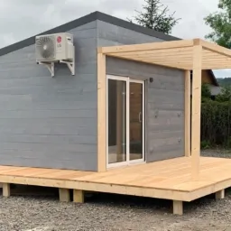 Szary domek szkieletowy z drewnianym tarasem i pergolą. Widoczny klimatyzator na ścianie. Ujęcie z poziomu gruntu, tło z zielenią i górami.