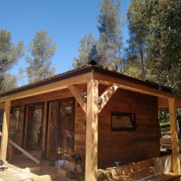 ŚwierG - Surowa drewniana altana w trakcie budowy, z widoczną konstrukcją dachu, deskami podłogowymi i elementami wykończeniowymi, otoczona zielenią drzew.