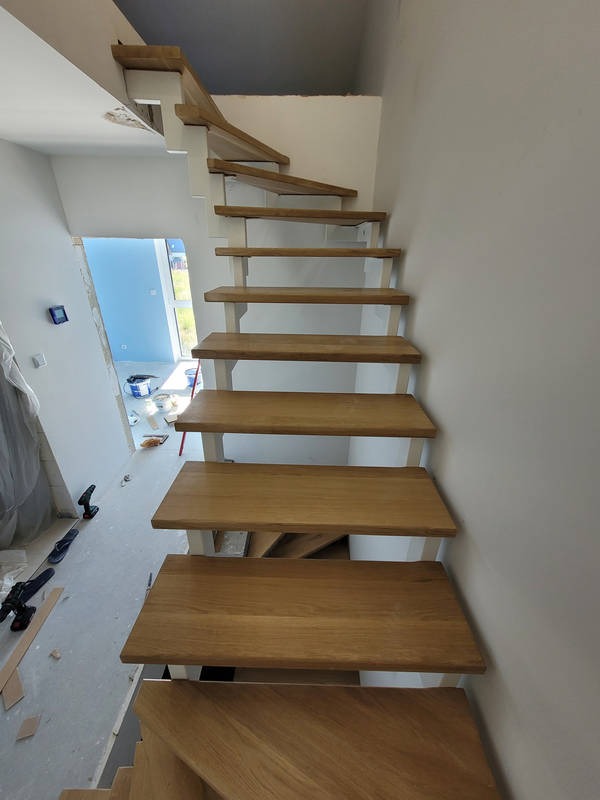 Nowoczesne, drewniane schody z białymi wspornikami w surowym wnętrzu. Widoczne narzędzia i materiały budowlane, podkreślające etap wykończenia.