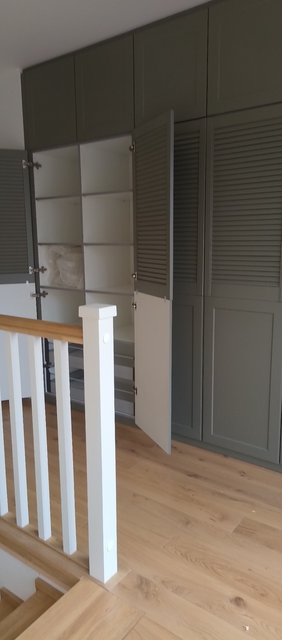 Szara szafa wnękowa z lamelowymi drzwiami, część otwartych, w tle fragment schodów z białą balustradą i drewniane podłogi. Widoczne półki i szuflady wewnątrz.