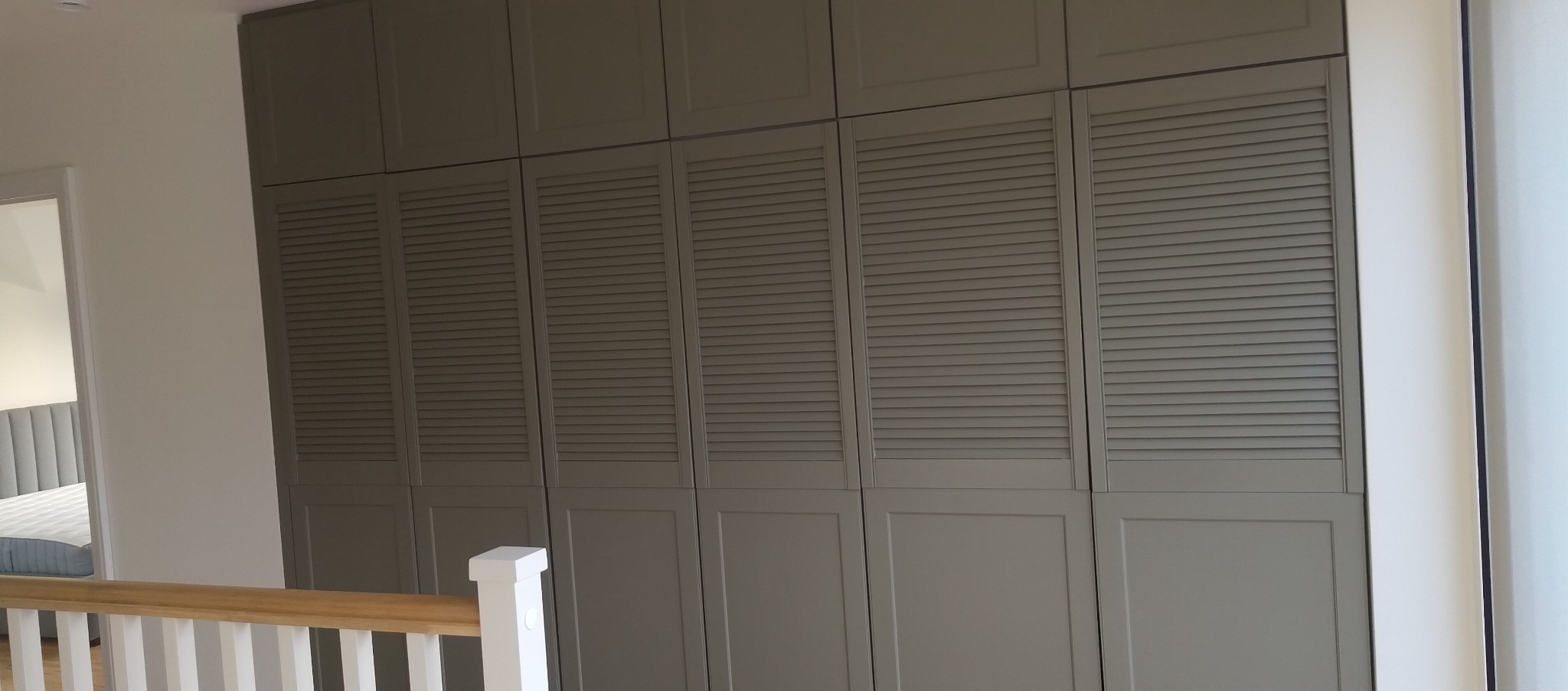 Szara szafa wnękowa z lamelowymi drzwiami, widoczna z perspektywy schodów. Fragment pokoju z łóżkiem w tle. Stonowana kolorystyka, minimalistyczny design.