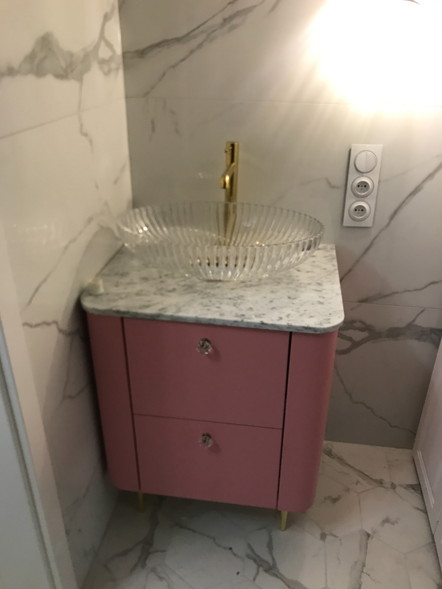 Różowa szafka łazienkowa z marmurowym blatem i szklaną umywalką, złota bateria, płytki imitujące marmur na ścianie i podłodze, nowoczesny styl.