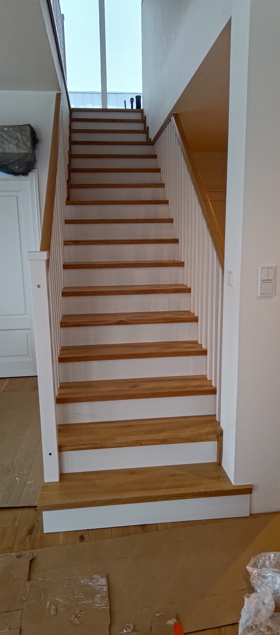 Drewniane schody z białymi balustradami w Krzywaczce, prowadzące na wyższą kondygnację. Widoczne zabezpieczenia podłogi na czas prac wykończeniowych.