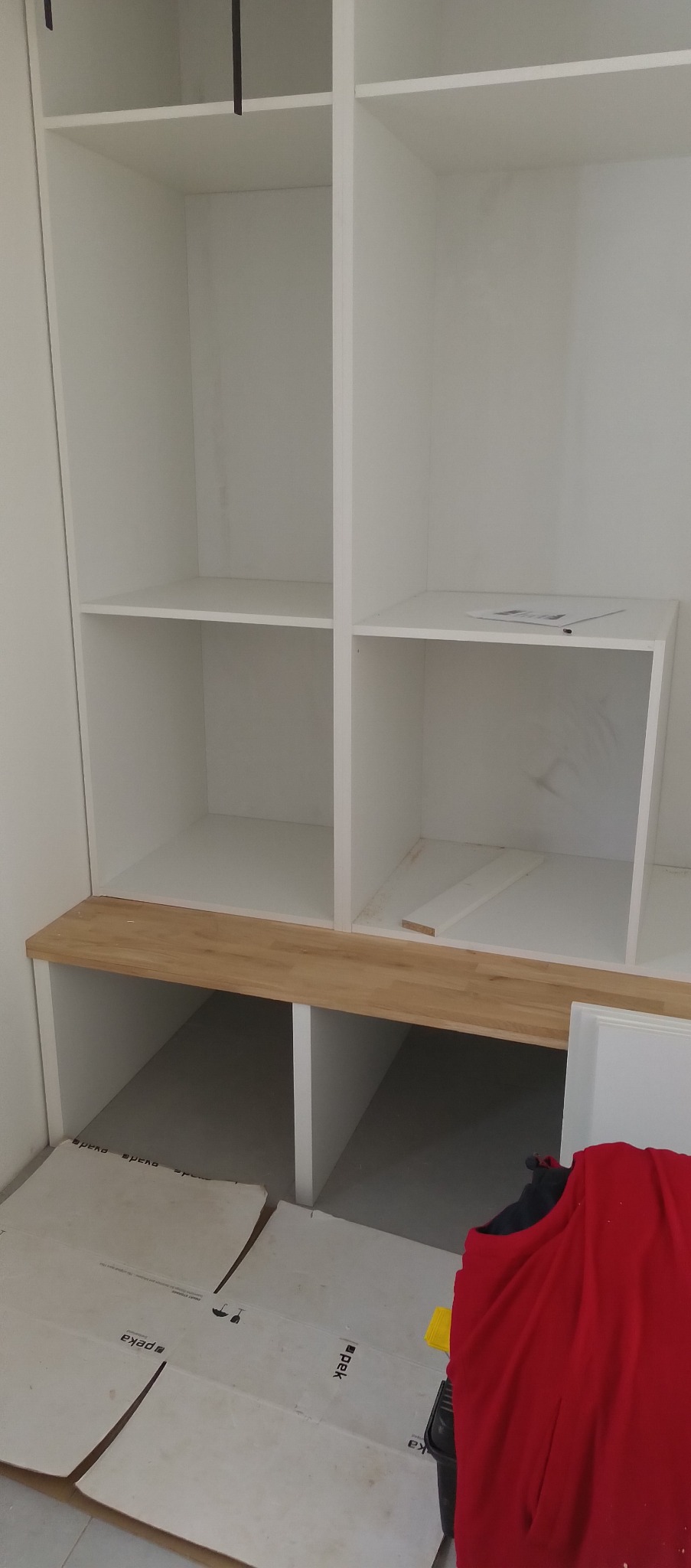 Wnętrze szafy wnękowej w trakcie montażu: białe półki, drewniany blat, kartonowe zabezpieczenie podłogi. Widoczne elementy wykończeniowe i narzędzia.