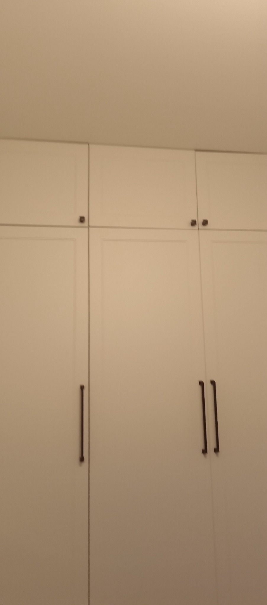 Biała szafa wnękowa z minimalistycznymi, długimi uchwytami. Górne szafki otwierane na dotyk. Prosty design, jasne wnętrze, pionowy kadr.