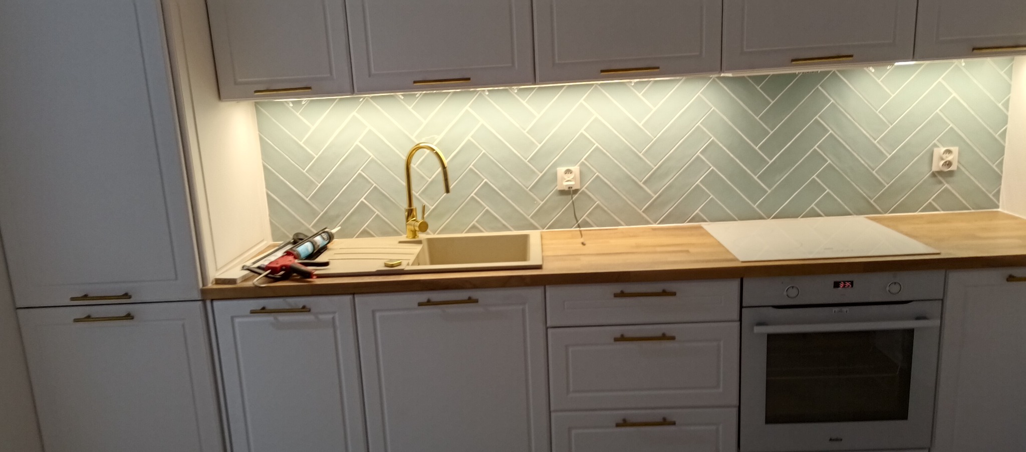 Biała kuchnia z złotymi akcentami: bateria, uchwyty. Na blacie widoczny silikon i pistolet do silikonu, co sugeruje montaż lub wykończenie. Ściana wyłożona kafelkami w jodełkę.