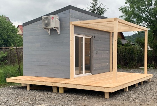 Szary domek szkieletowy z drewnianym tarasem i pergolą. Widoczny klimatyzator na ścianie. Ujęcie z poziomu gruntu, tło z zielenią i górami.