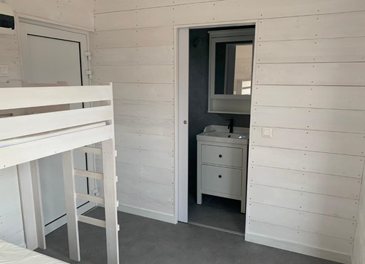 Jasne wnętrze domku szkieletowego z drewnianą boazerią, łóżkiem piętrowym i łazienką z umywalką, widoczne przez otwarte drzwi przesuwne.