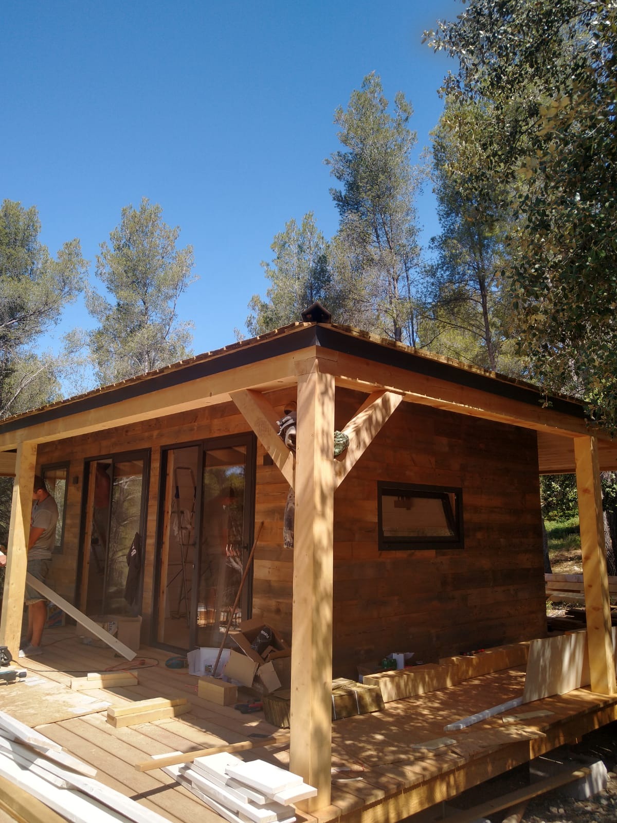 Surowa drewniana altana w trakcie budowy, z widoczną konstrukcją dachu, deskami podłogowymi i elementami wykończeniowymi, otoczona zielenią drzew.