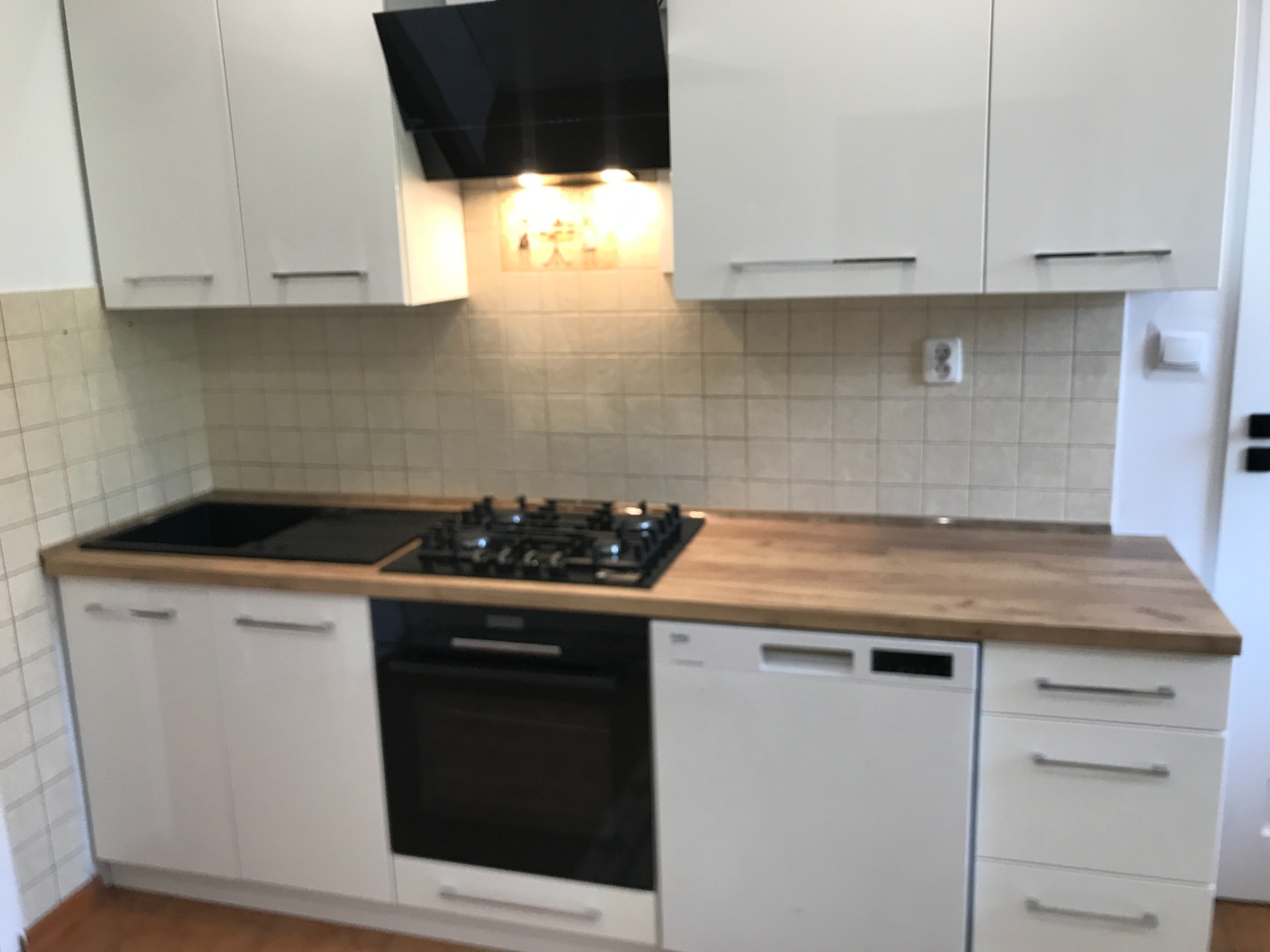 Biała zabudowa kuchenna z drewnianym blatem, płyta gazowa i piekarnik w zabudowie. Widoczne górne szafki i okap. Jasne płytki na ścianie.