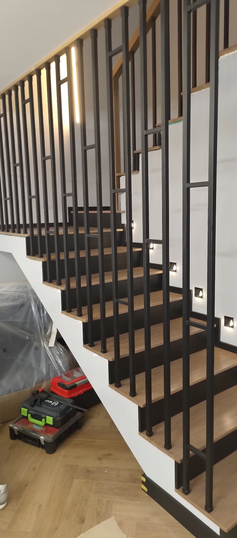 Nowoczesne schody drewniane z czarną, metalową balustradą i subtelnym oświetleniem punktowym w ścianie. Widoczne narzędzia sugerują etap wykończeniowy.