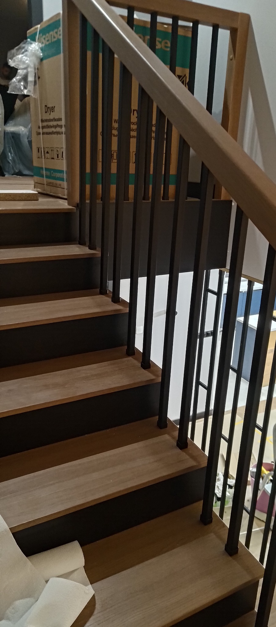 Nowoczesne schody z drewnianymi stopniami i czarnymi balustradami. Ujęcie z dołu, widoczne detale wykończenia i kontrast kolorów. Częściowo widoczny karton w tle.