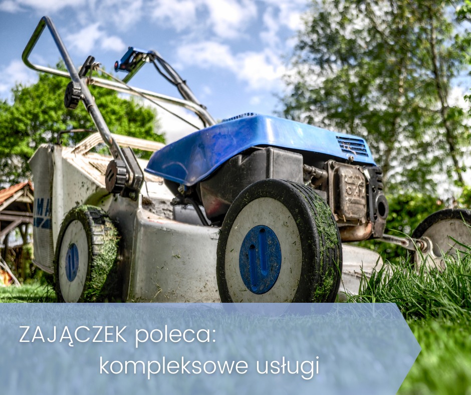 Używana kosiarka spalinowa z niebieską obudową silnika, z resztkami trawy na kołach, stojąca na trawniku, z widocznym budynkiem w tle i logotypem firmy na pierwszym planie.