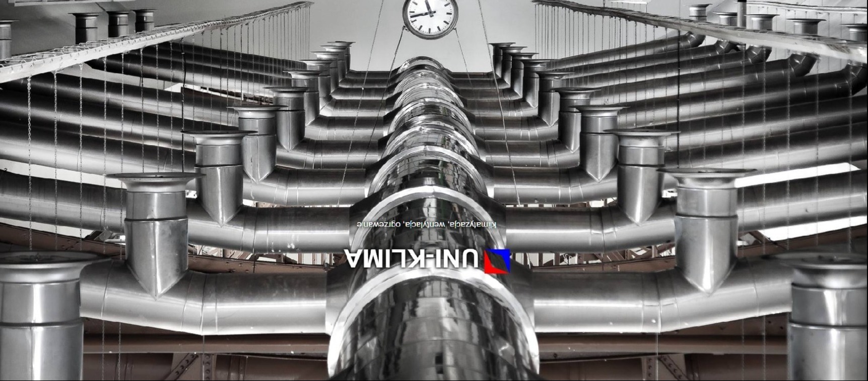 Srebrne, metalowe rury wentylacyjne podwieszone pod sufitem, widok z dołu, z widocznym logo firmy Uni-Klima i zegarem na ścianie w tle.