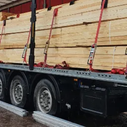 Ciężarówka z naczepą przewozi ładunek świeżo przetartego drewna, zabezpieczonego czerwonymi pasami transportowymi, widoczne koła i błotniste podłoże.