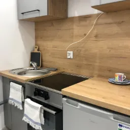 Kompaktowa kuchnia w szaro-drewnianej tonacji z elementami wyposażenia: zlew, płyta grzewcza, piekarnik, mała lodówka, drewniana deska do krojenia i kubek na blacie.