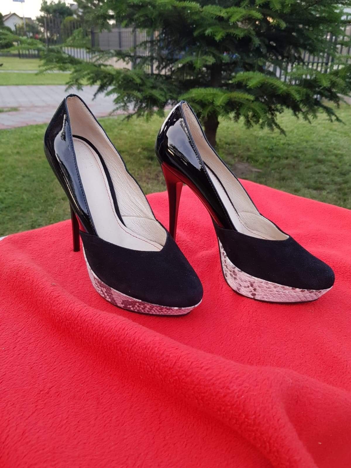 Para czarno-lakierowanych szpilek z zamszowymi wstawkami i platformą imitującą skórę węża, ustawiona na czerwonej tkaninie, z drzewem w tle.