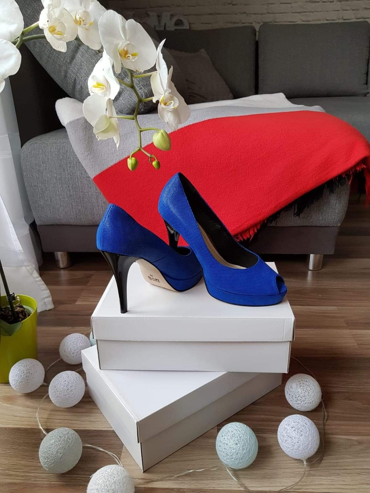 Kobaltowe szpilki z odkrytymi palcami, ustawione na białych pudełkach, z dekoracyjną girlandą świetlną i orchideą w tle, na tle szarej sofy z czerwonym pledem.