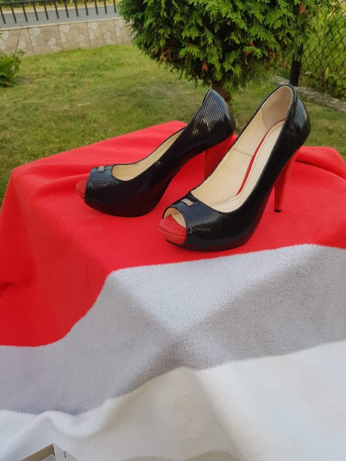Para czarnych lakierowanych szpilek z czerwonymi obcasami i otwartymi palcami, ustawiona na czerwono-białym kocu w plenerze.
