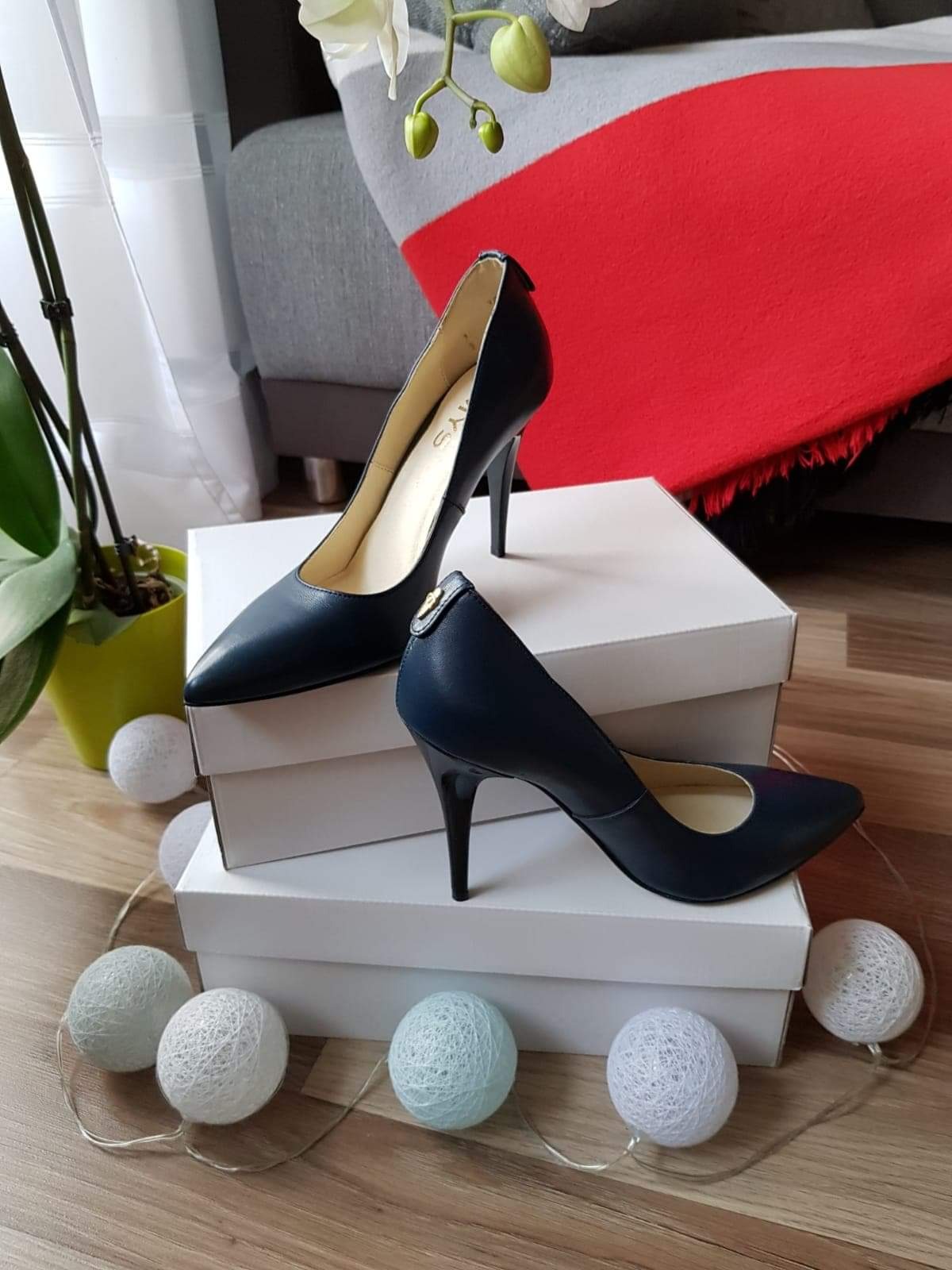 Para granatowych skórzanych szpilek na białych pudełkach, otoczona dekoracyjnymi kulami świetlnymi, z rośliną w tle.