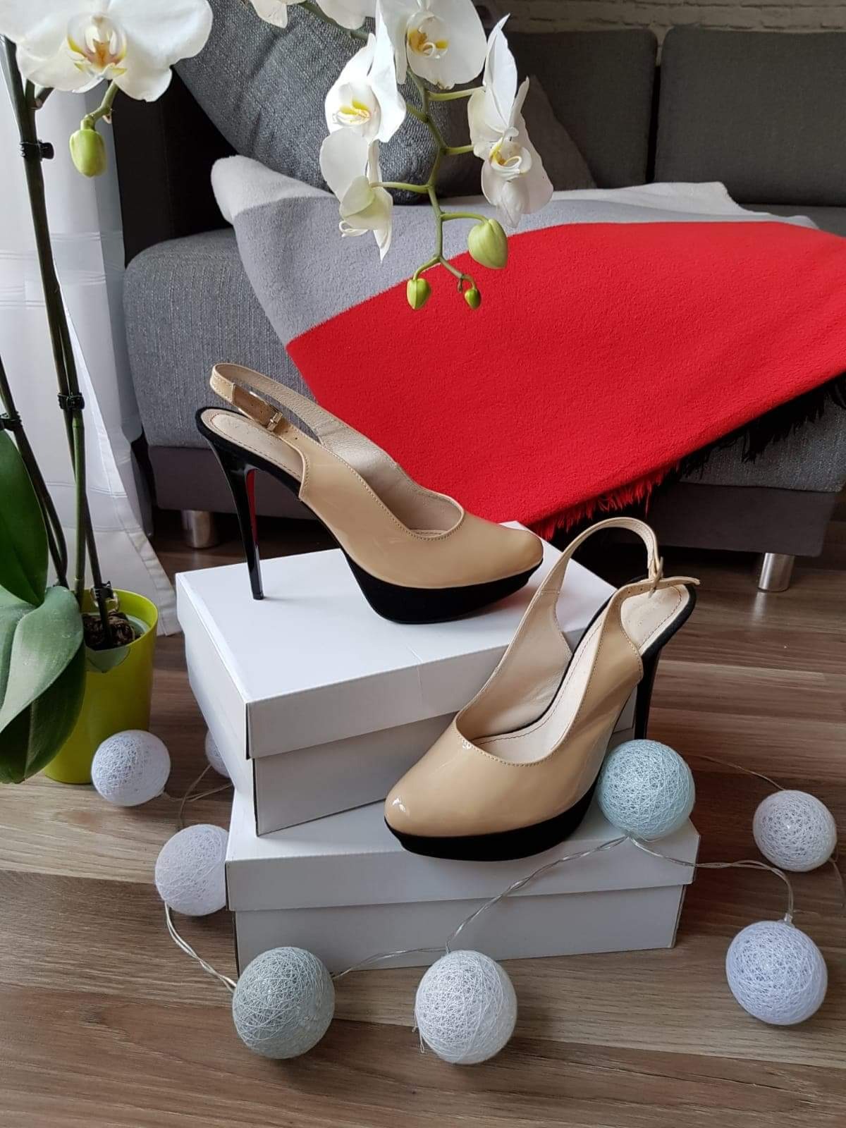 Beżowe lakierowane czółenka na platformie z czarnym obcasem typu szpilka, ustawione na białych pudełkach, otoczone dekoracyjnymi kulami świetlnymi, z orchideą i szarym narożnikiem w tle.