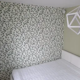 Pokój z łóżkiem, ściana pokryta tapetą w liście eukaliptusa, nowoczesna geometryczna lampa sufitowa i białe reflektory punktowe.