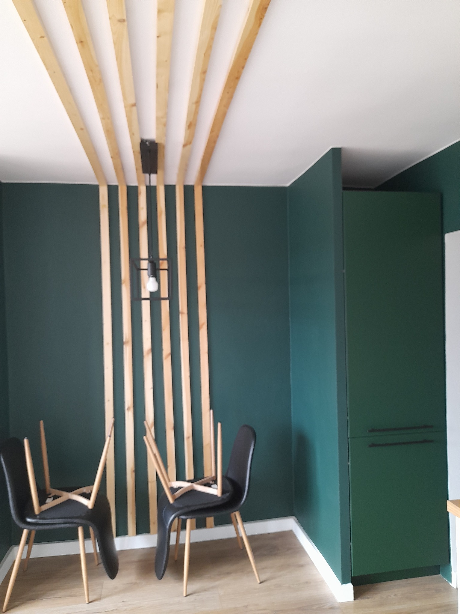 Wnętrze z ciemnozielonymi ścianami, drewnianymi listwami na suficie i za lampą, dwie czarne krzesła odwrócone do góry nogami, zielona szafka w rogu.
