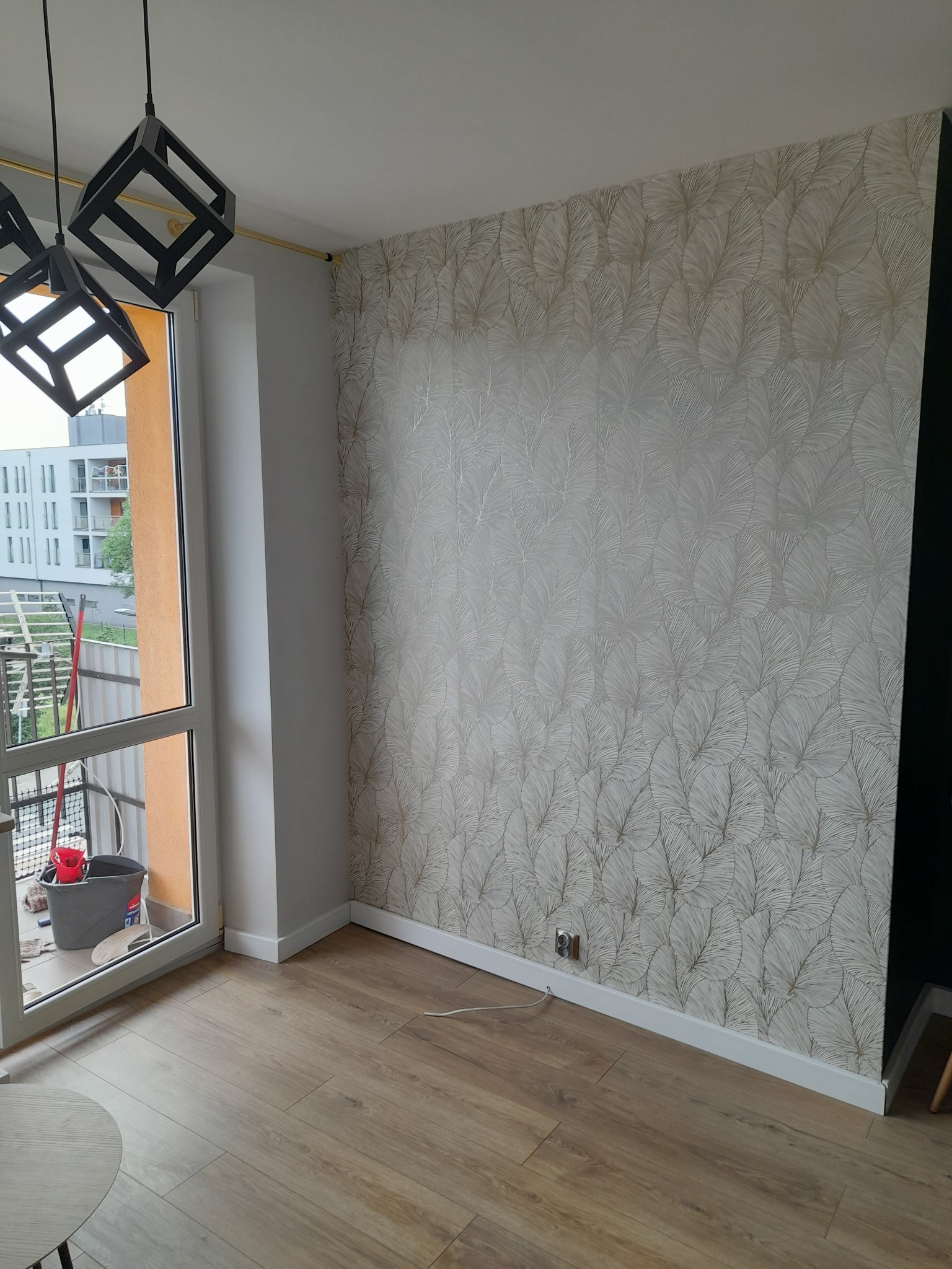 Pokój po remoncie: ściana z tapetą w liście, drewniana podłoga, nowoczesne lampy wiszące, widok na balkon z narzędziami.