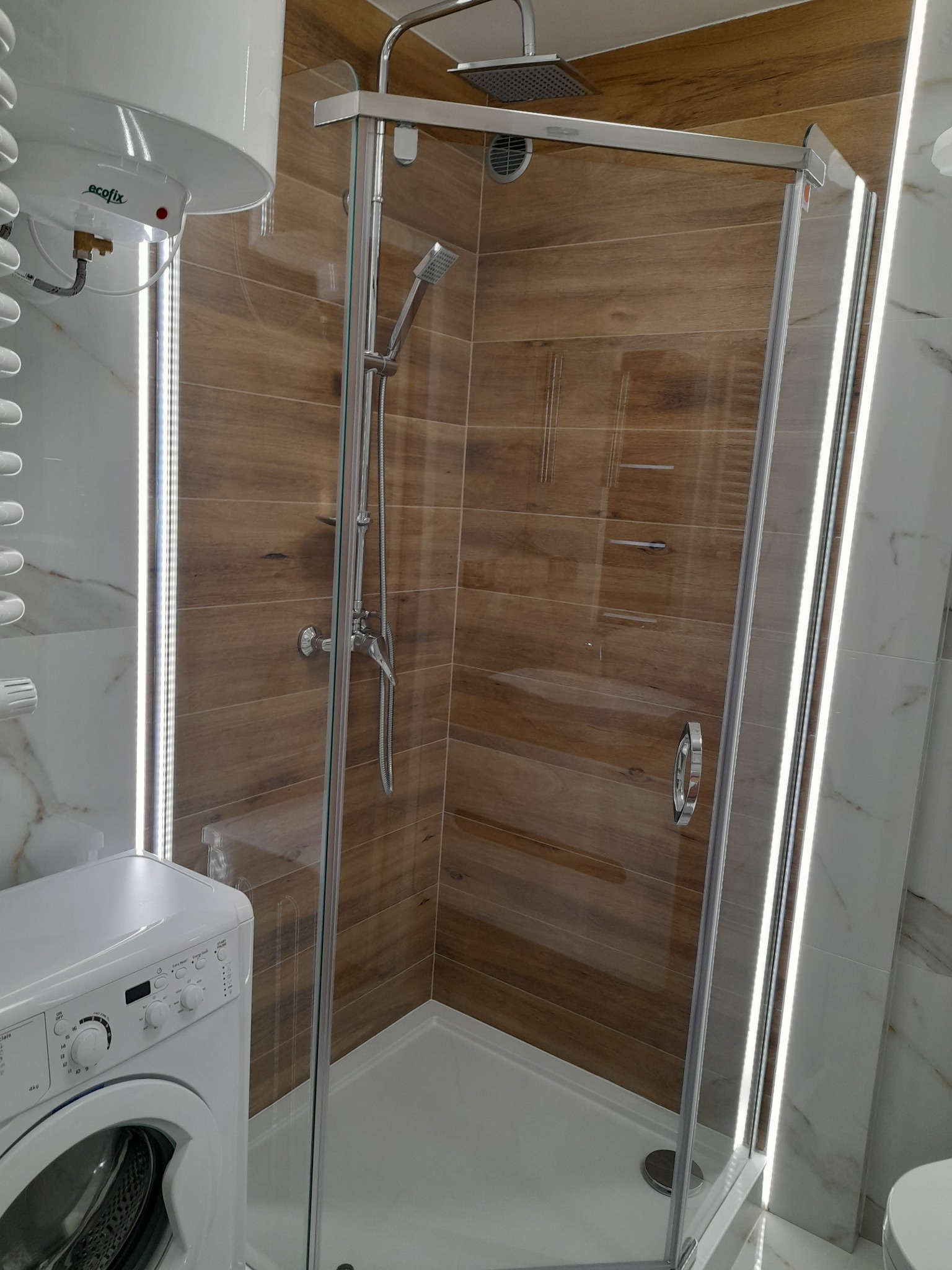 Nowoczesna kabina prysznicowa z drewnopodobnymi płytkami i oświetleniem LED, widoczna pralka i bojler.