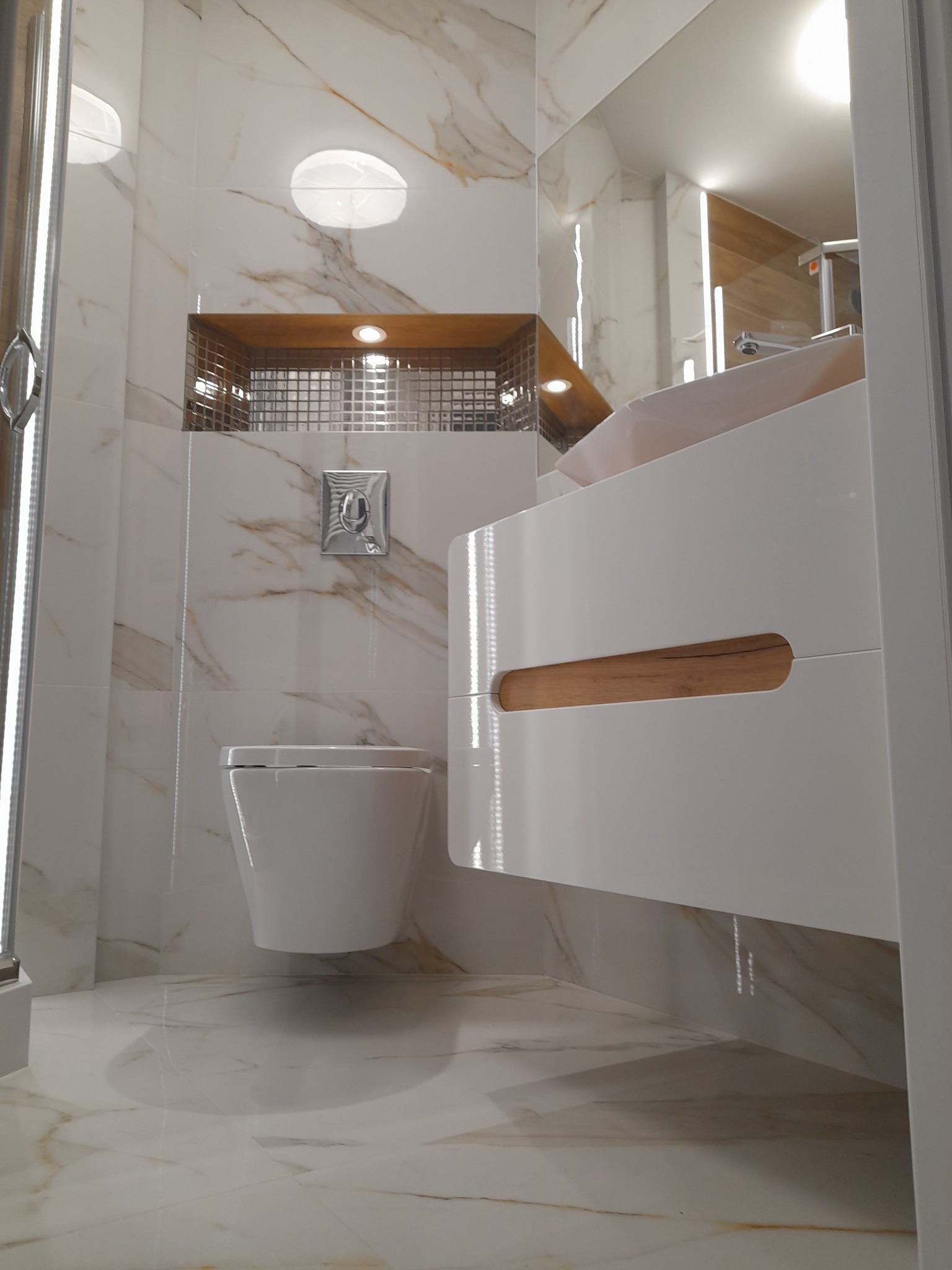 Nowoczesna łazienka z marmurowymi płytkami, podwieszaną toaletą, białą szafką umywalkową z drewnianym akcentem i wnęką z mozaiką, oświetlona punktowo.