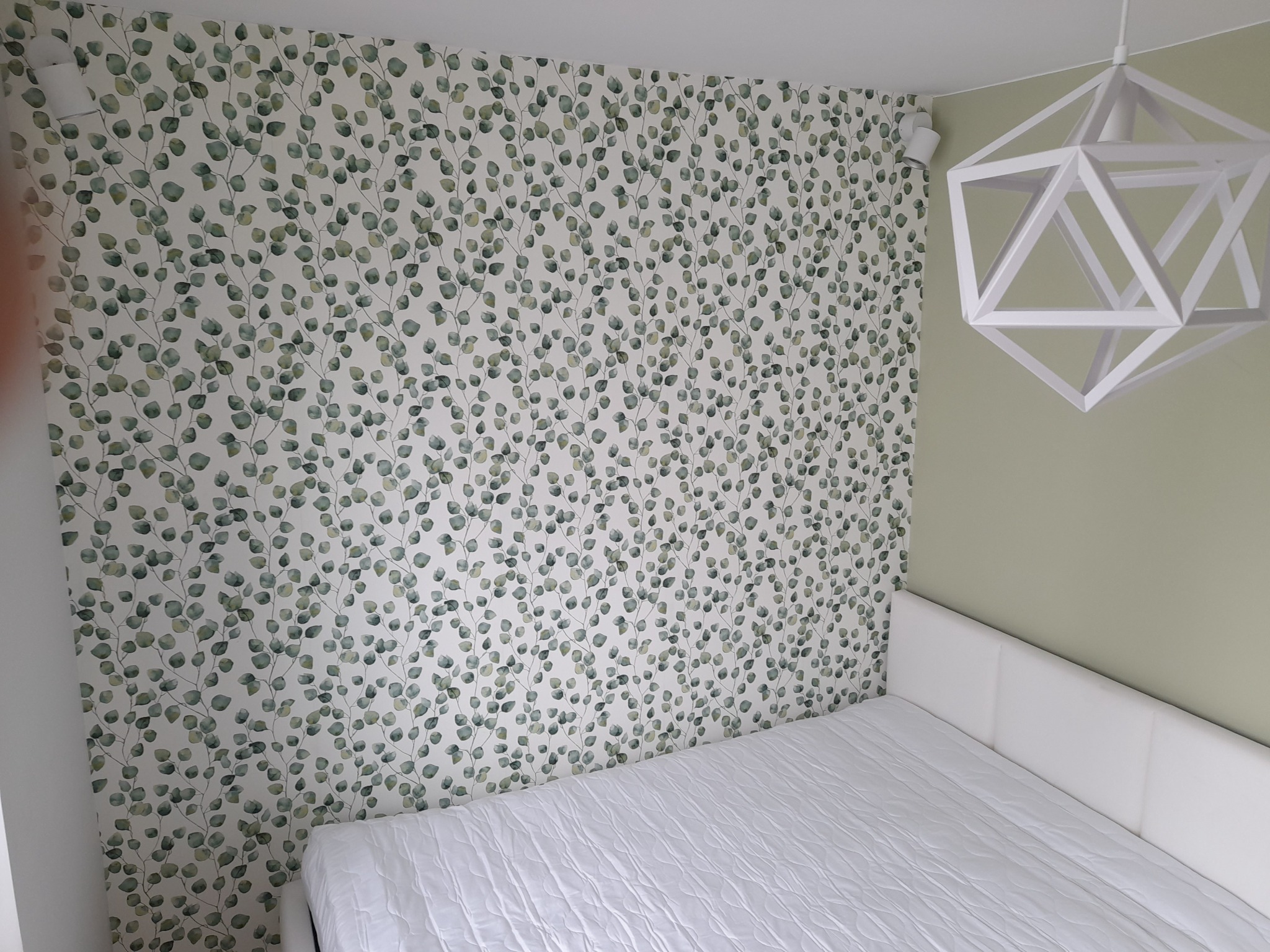 Pokój z łóżkiem, ściana pokryta tapetą w liście eukaliptusa, nowoczesna geometryczna lampa sufitowa i białe reflektory punktowe.