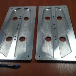 Dwie aluminiowe płyty z wyfrezowanymi wgłębieniami i otworami, leżące na drewnianym stole, z długopisem u dołu kadru, ukazujące precyzyjną obróbkę metalu.