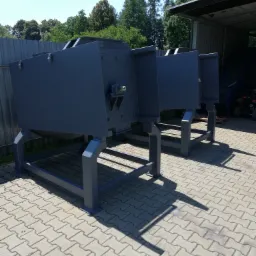 Szare metalowe konstrukcje na kostce brukowej, prawdopodobnie elementy większej maszyny, w tle drzewa i zadaszona hala.