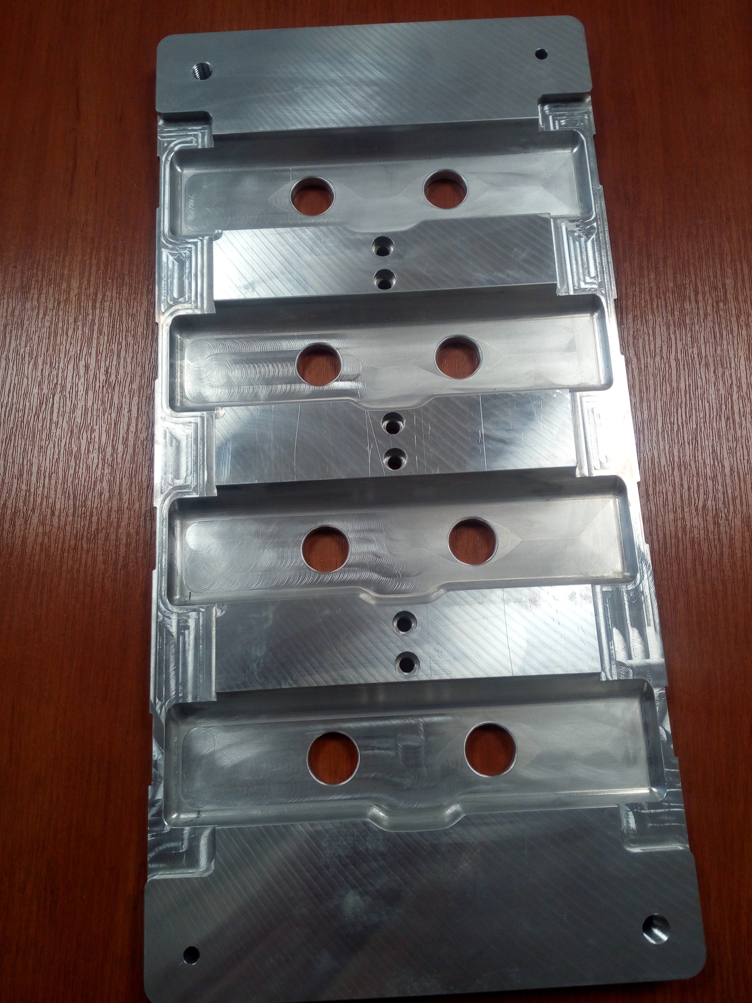 Precyzyjnie obrobiony, srebrny, metalowy element z czterema wgłębieniami i okrągłymi otworami, leżący na drewnianej powierzchni, widoczne ślady obróbki skrawaniem.