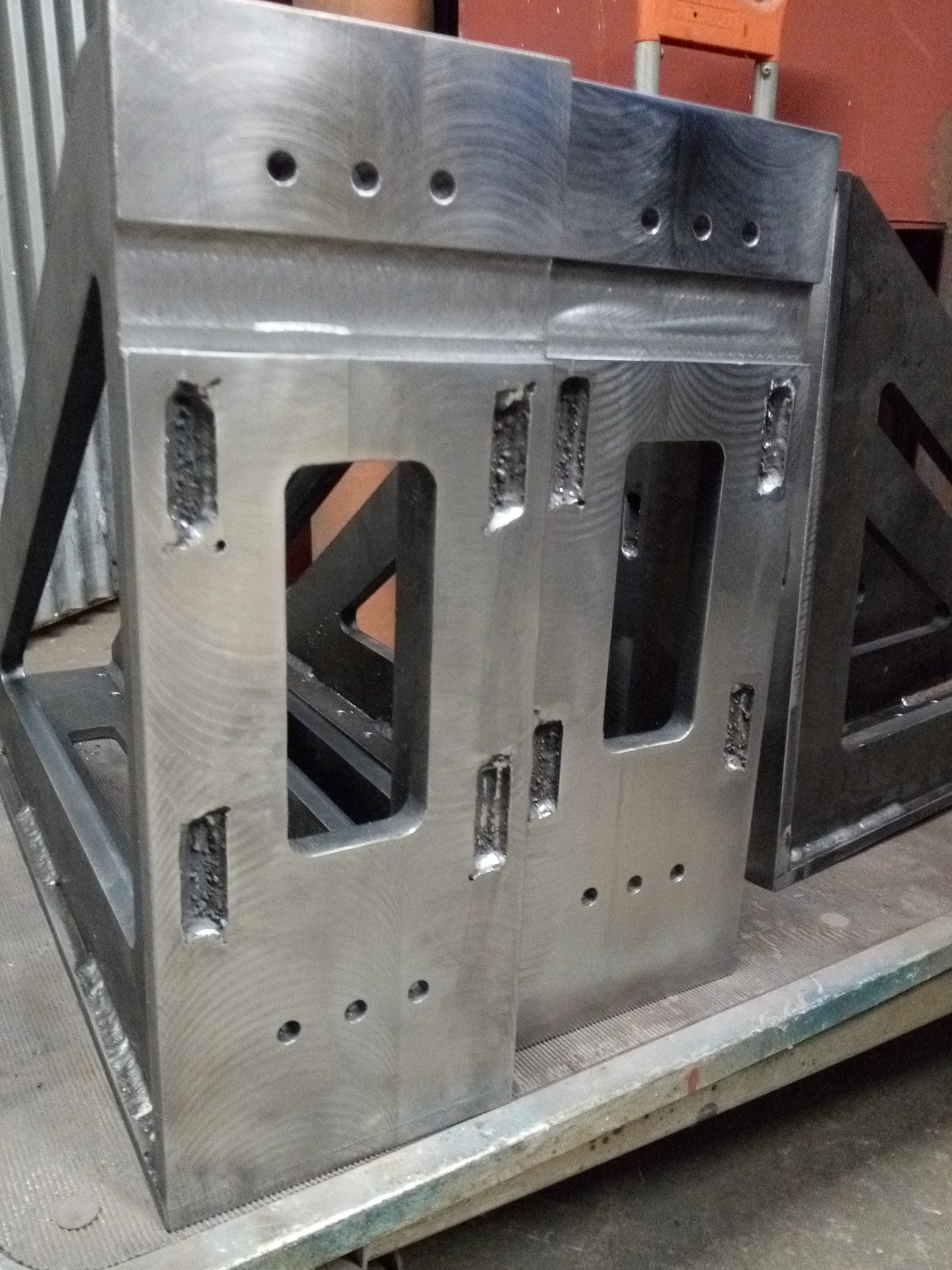 Dwa szare, metalowe elementy konstrukcyjne z wyciętymi otworami i widocznymi śladami obróbki, ustawione obok siebie na metalowej powierzchni.