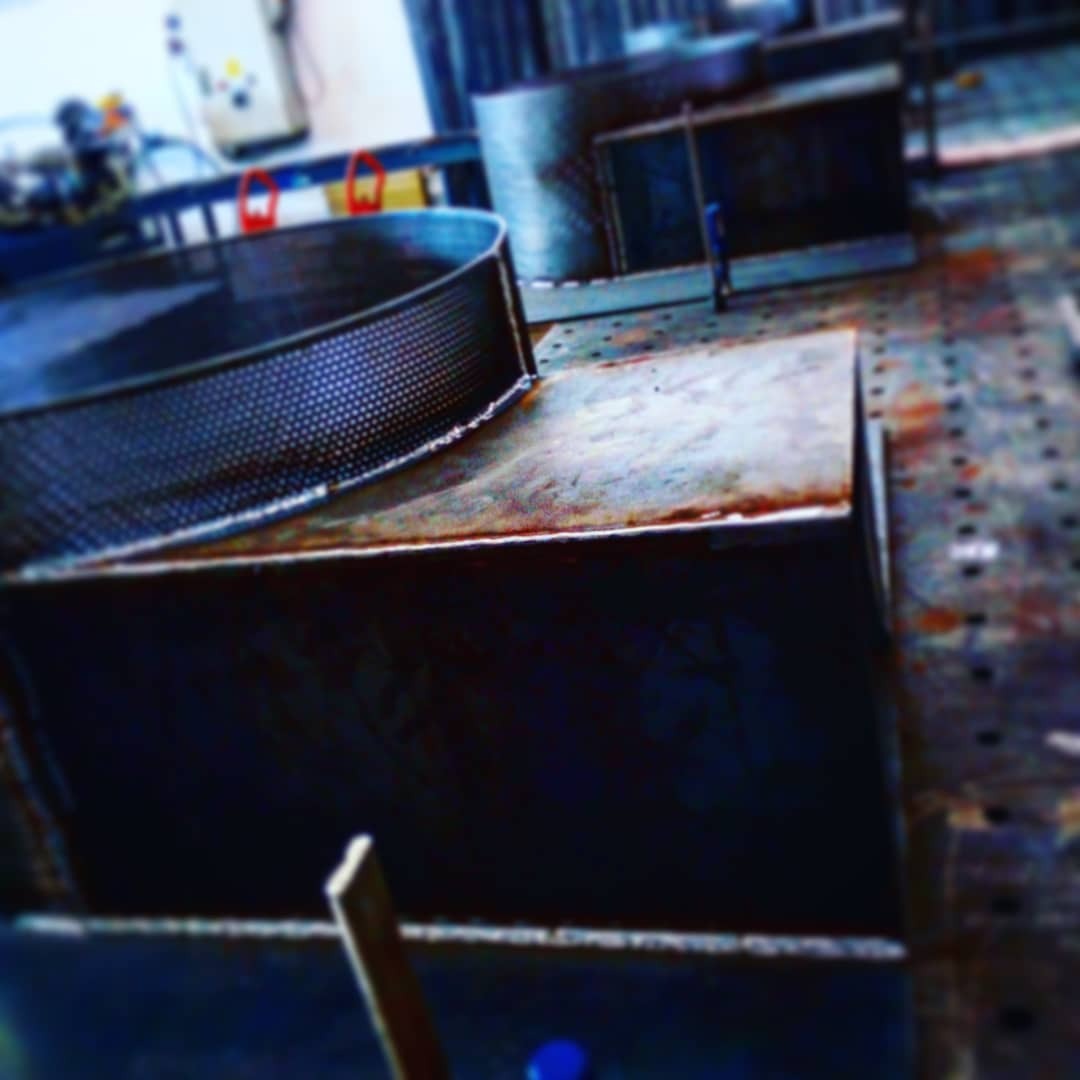 Metalowe elementy konstrukcyjne, w tym perforowany pierścień i prostopadłościenne kształtowniki, ułożone na perforowanym stole spawalniczym w warsztacie.