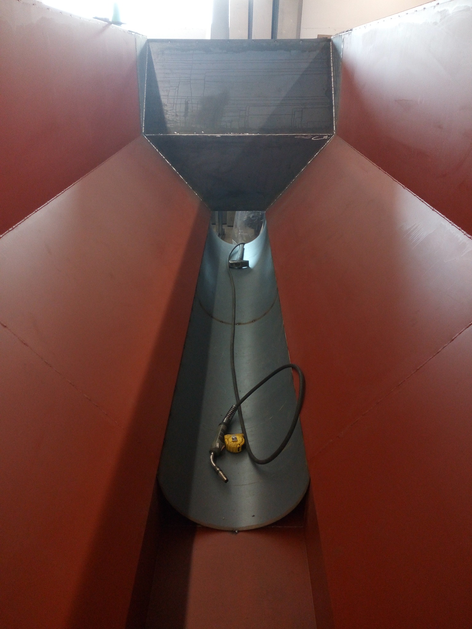 Wnętrze konstrukcji stalowej w kształcie stożka, widok z góry, z widocznym uchwytem spawalniczym na dnie, sugerujące etap spawania lub kontroli po spawaniu.