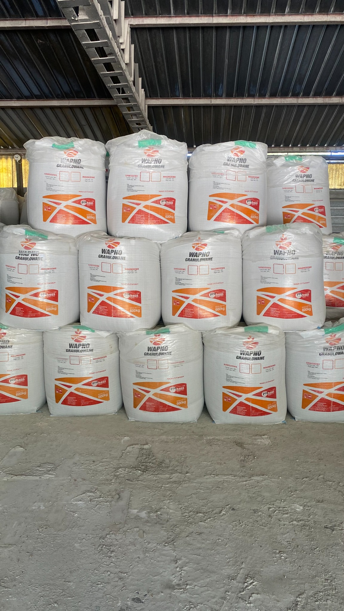 Stos palet z workami wapna granulowanego w magazynie z blachy falistej, widoczne etykiety producenta i informacje o wadze 600kg.