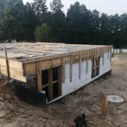 Budowa domu jednorodzinnego w stanie surowym: fundamenty z bloczków betonowych, szalunki drewniane, hydroizolacja z folii kubełkowej, widok na las i jezioro w tle, pies w prawym dolnym rogu.