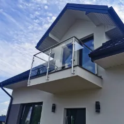 Nowoczesny balkon z balustradą szklaną i metalowymi słupkami, widok z dołu na tle błękitnego nieba z obłokami, elewacja domu w jasnych kolorach.